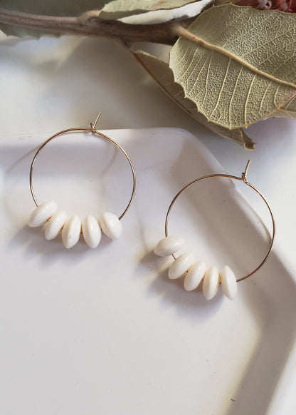 A pair of white bone bead hoop earrings with gold-filled hoops, displayed on a white surface with some green leaves in the background.